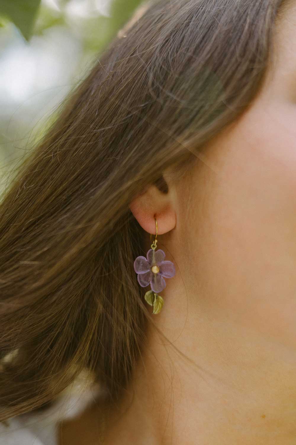 Close-up of a person wearing a purple flower-shaped handcrafted earring with a small green leaf detail, inspired by Impatiens jewelry, as brown hair partially covers their ear—perfect for lovers of nature-inspired accessories.