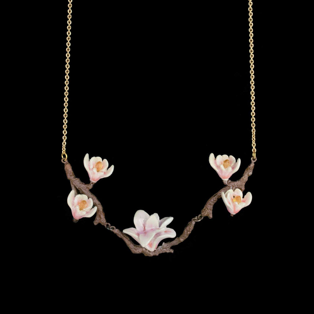 A delicate necklace featuring a curved brown branch pendant adorned with pink and white magnolia blossoms, suspended from a fine gold chain against a solid black background.