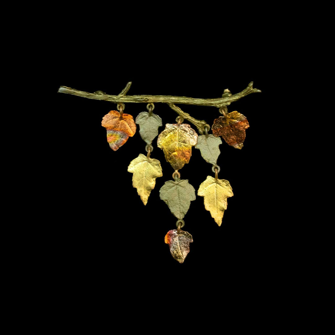 A decorative branch-shaped ornament or brooch with dangling leaf charms in metallic gold, olive green, and coppery orange tones, shown against a solid black background.