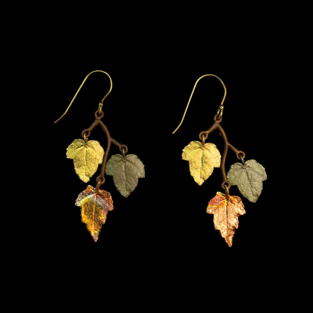 A pair of dangling earrings shaped like autumn leaves on thin brown branches, with gold fishhook ear wires. The leaves are textured and show shades of yellow, olive green, coppery orange, and brown, set against a solid black background.