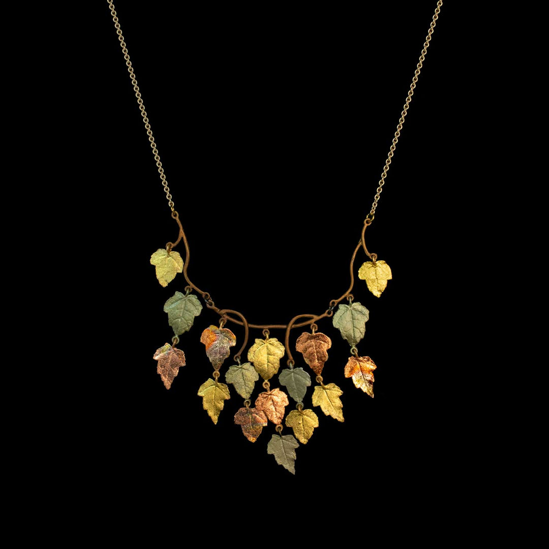A delicate necklace featuring multiple small leaf-shaped charms in yellow, green, and copper tones hanging from a gold-toned chain, set against a solid black background.