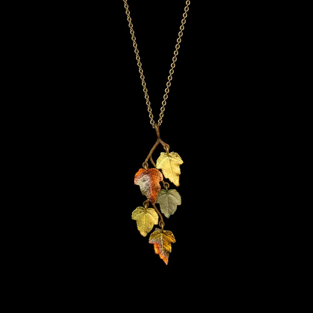 A gold chain necklace featuring a pendant shaped like a small branch with several textured, metallic autumn leaves in yellow, orange, and olive green, set against a solid black background.