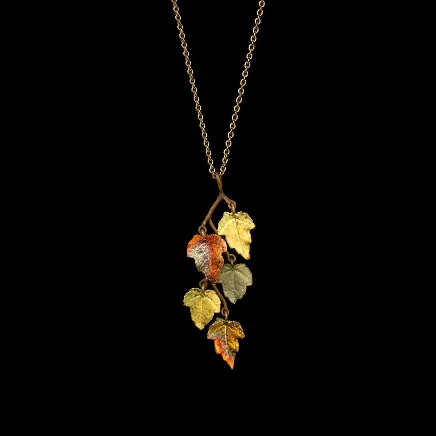 A gold chain necklace featuring a pendant shaped like a small branch with several textured, metallic autumn leaves in yellow, orange, and olive green, set against a solid black background.