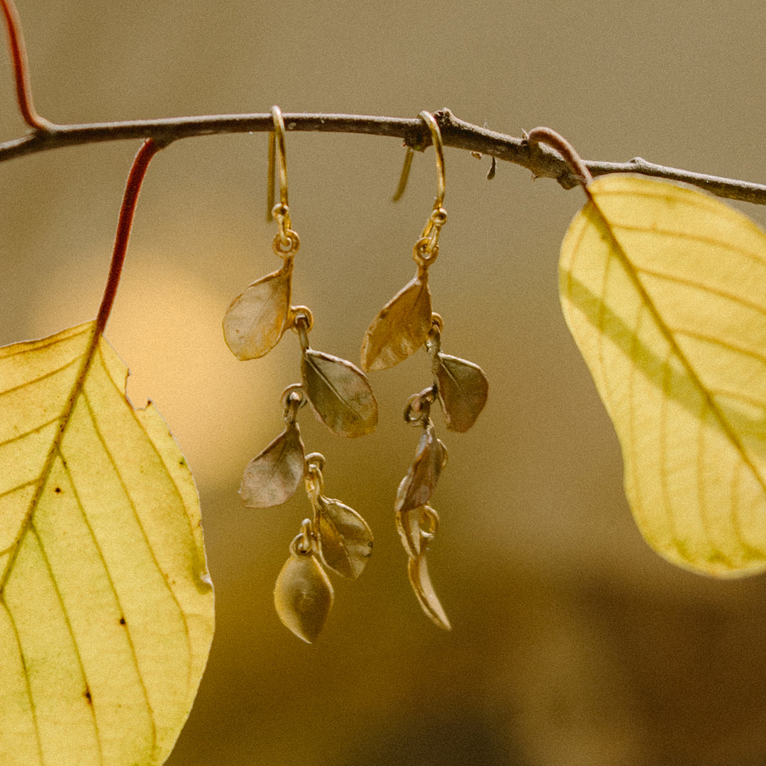 Gold and orange earrings with leaf-shaped pendants hang from a thin branch, surrounded by yellow leaves against a blurred background.