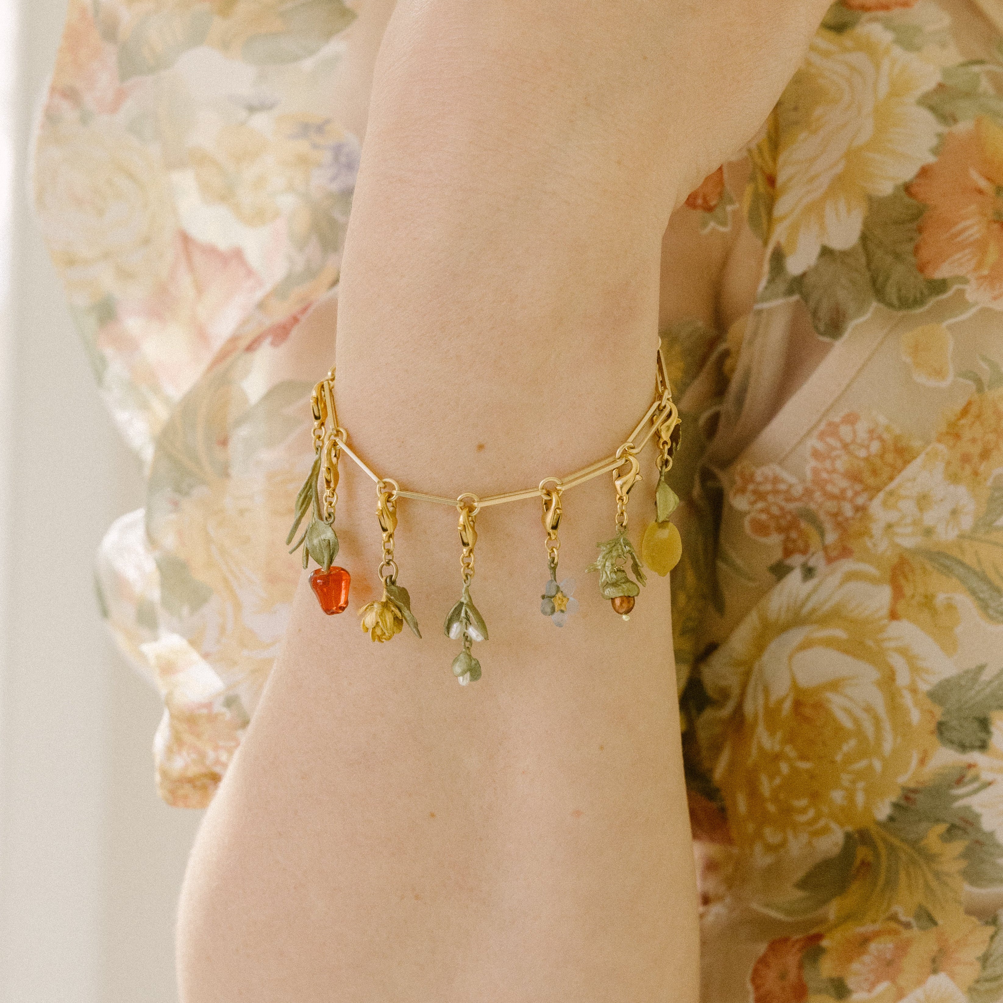 A person wearing a gold charm bracelet with small flower and fruit charms, paired with a sheer, floral-patterned blouse and an extender for adjustable jewelry styling.